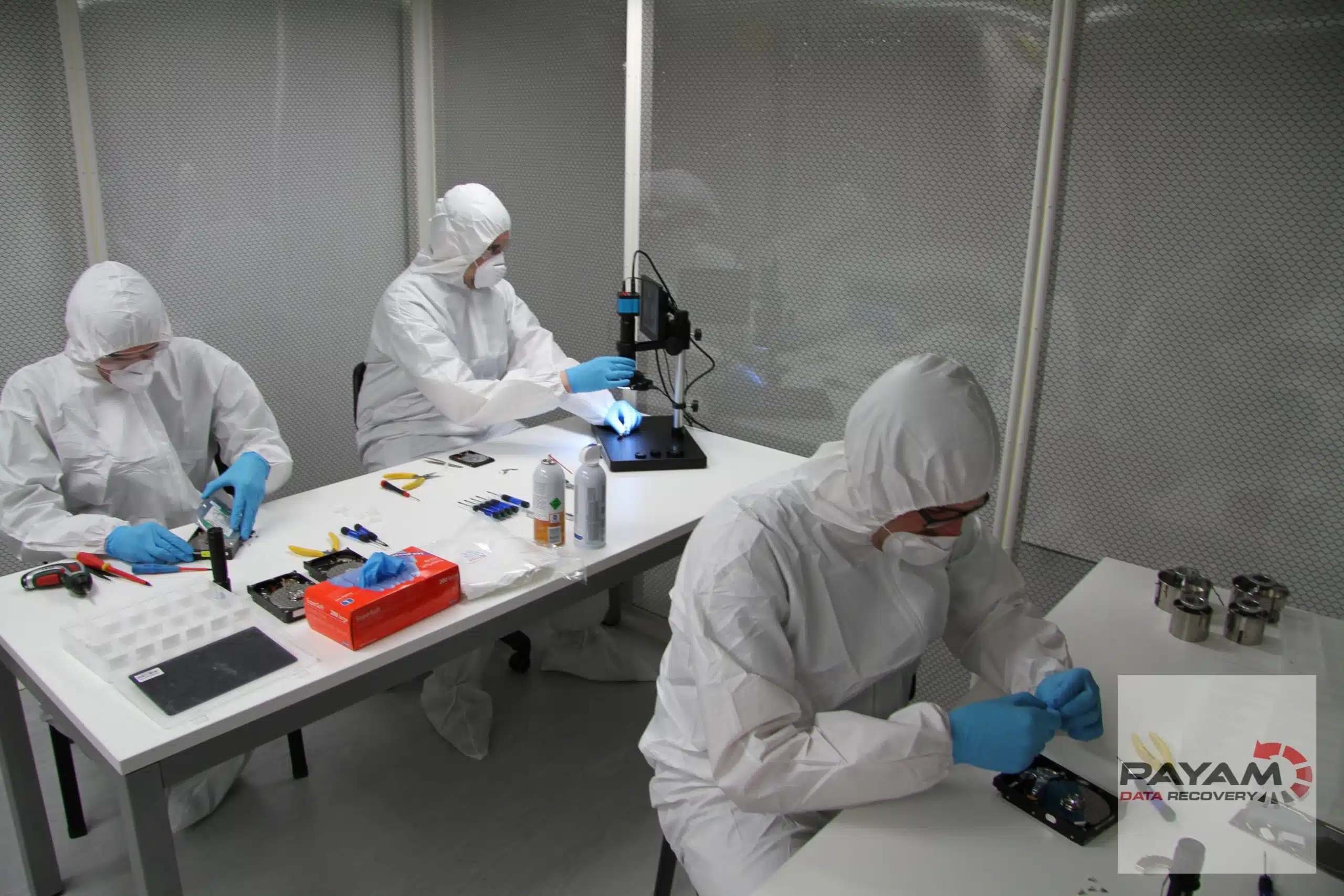 Three technicians in white cleanroom suits working at a table under a laminar flow hood
